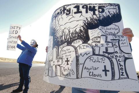 Two protesters holding signs with grave markers and image of nuclear bomb cloud stand at side of the road. 