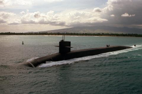 Image of large black nuclear submarine in centre of photograph, travelling on surface of sea. There is a coast line behind and a mountain as well as a cloudy sky in background. 