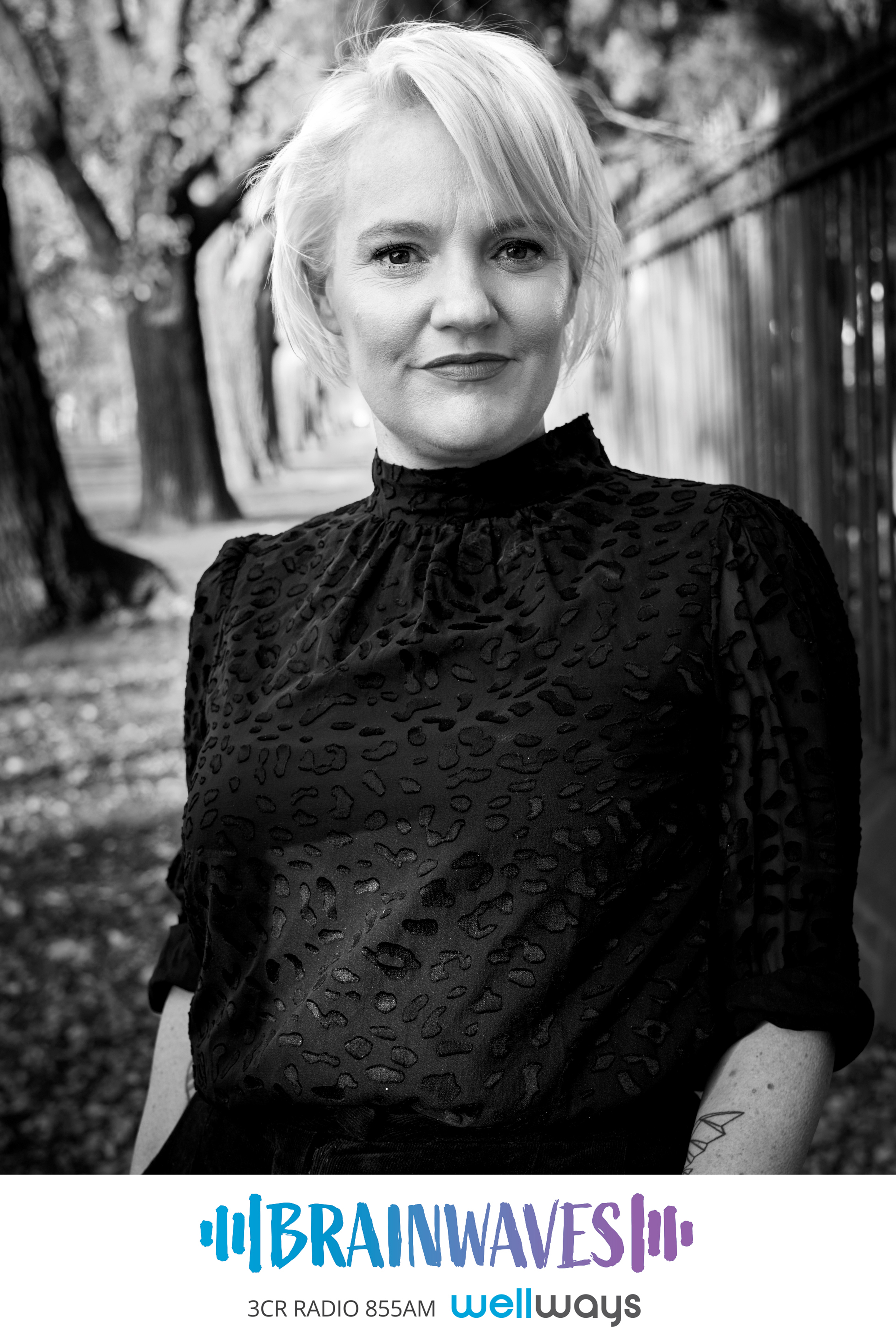 Jacinta Parsons is a caucasian woman with short platinum blonde hair, she wears a black lacy short sleeved top and black trousers and dark lipstick, the photo is black and white, in the image she is standing outside in a park with trees behind her as she smiles at the camera.