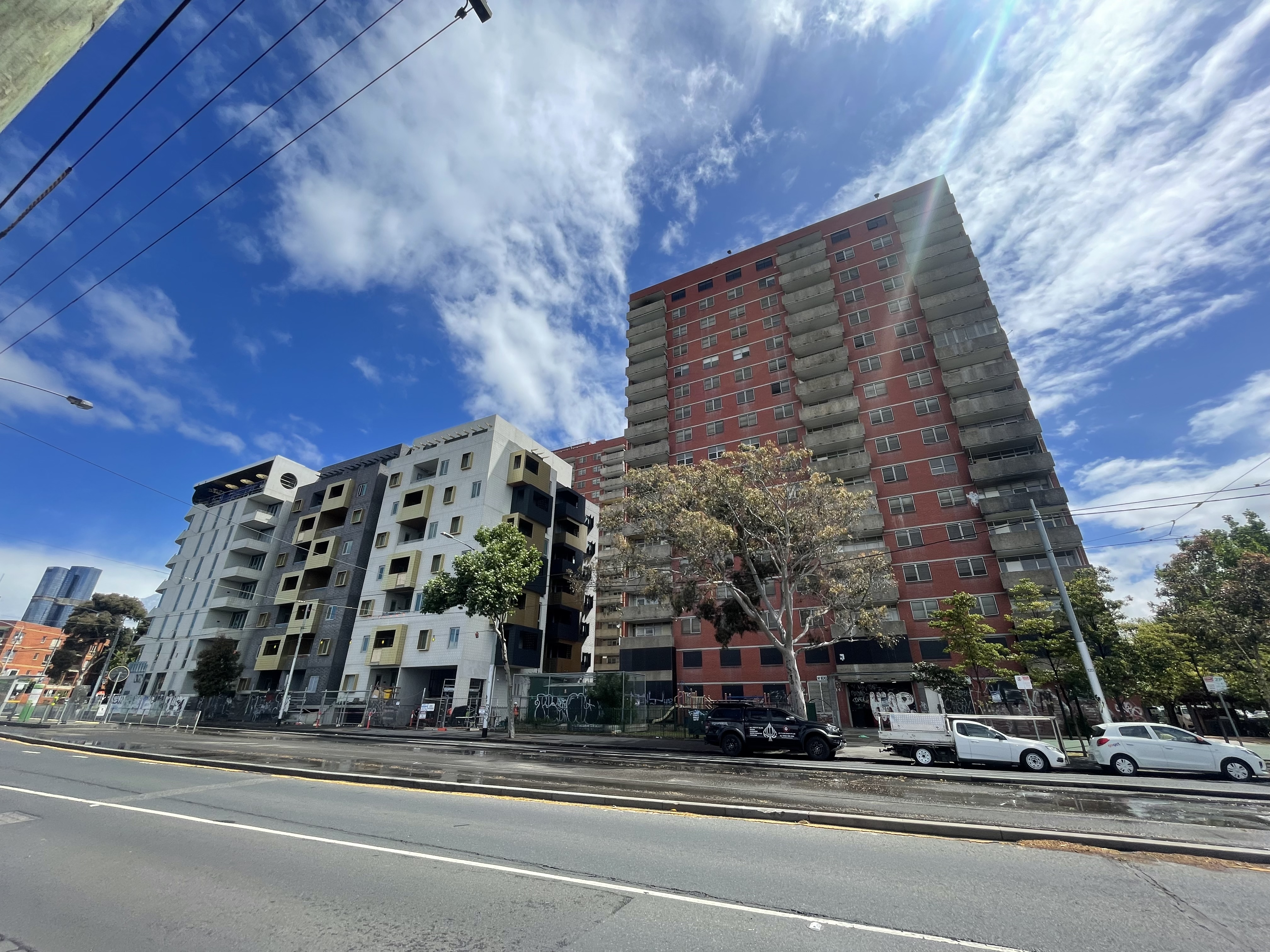 New ‘community’ housing currently being built alongside the decommissioned public housing estate at 141 Nicholson St, Carlton. The new building on the left is short with stainless steel cladding and no green space, and the boarded up 16-storey brick building on the right has balconies and graffiti on the base of its façade. The sky is cloudy and traffic is passing on a street in the foreground with tram tracks and multiple lanes.