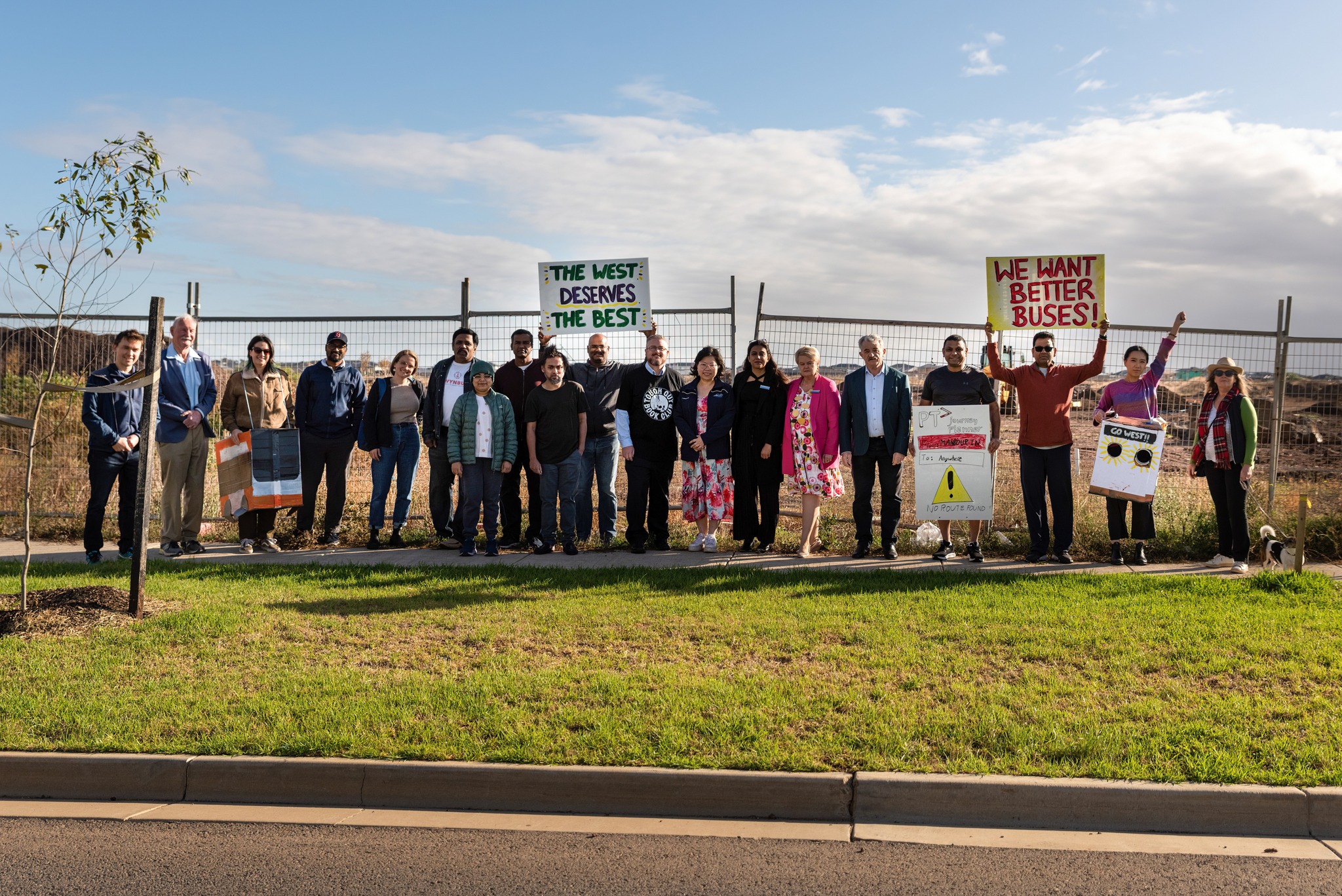 A group of people wearing colourful clothing standing in the sun in front of an empty demolition site holding placards that read "the west deserves the best", "we want better buses!" and other signs too small to read.