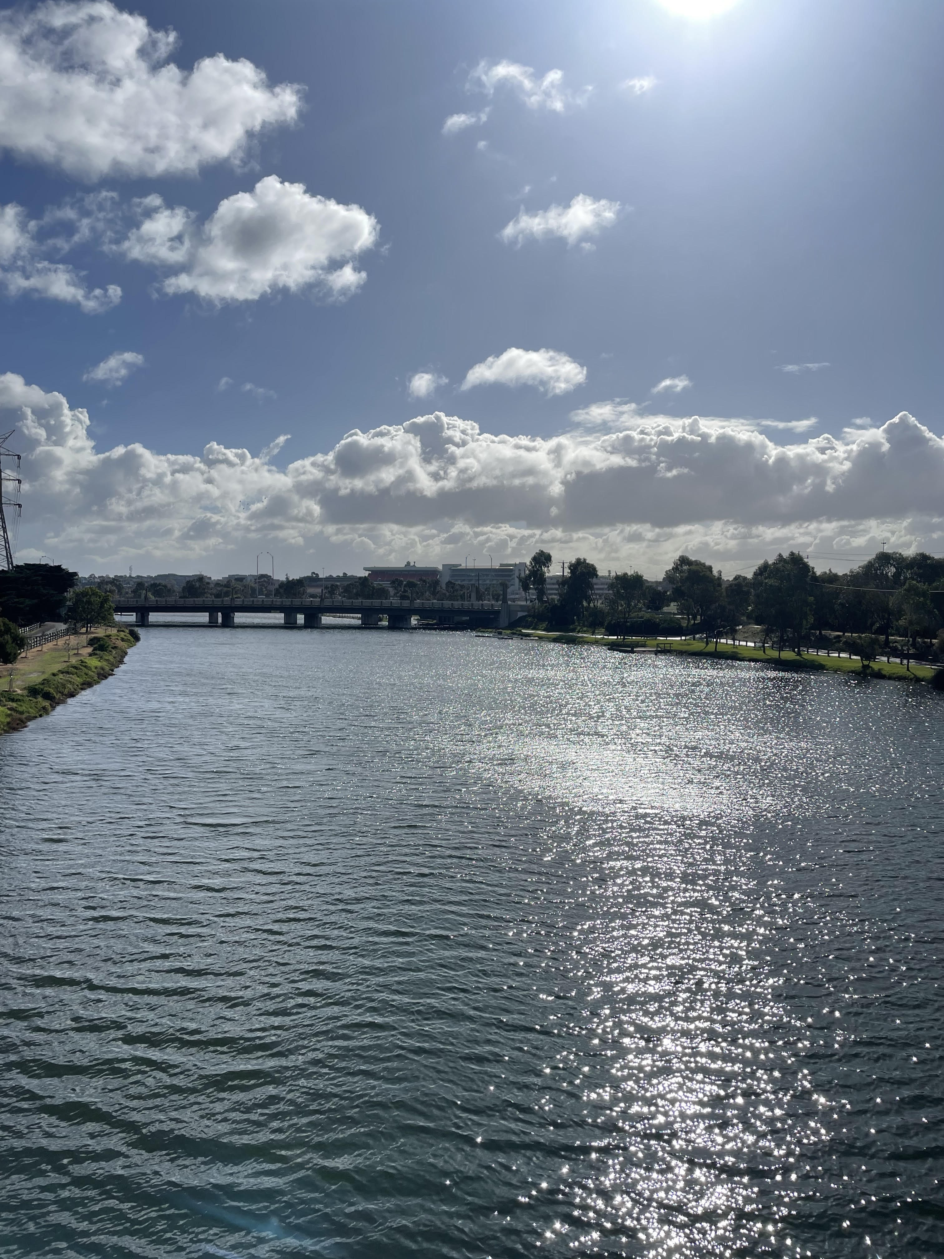 A photograph of the Maribyrnong River, on a sunny and partly cloudy day, with the sun reflecting on its surface, a bridge in the distance and trees on either side.