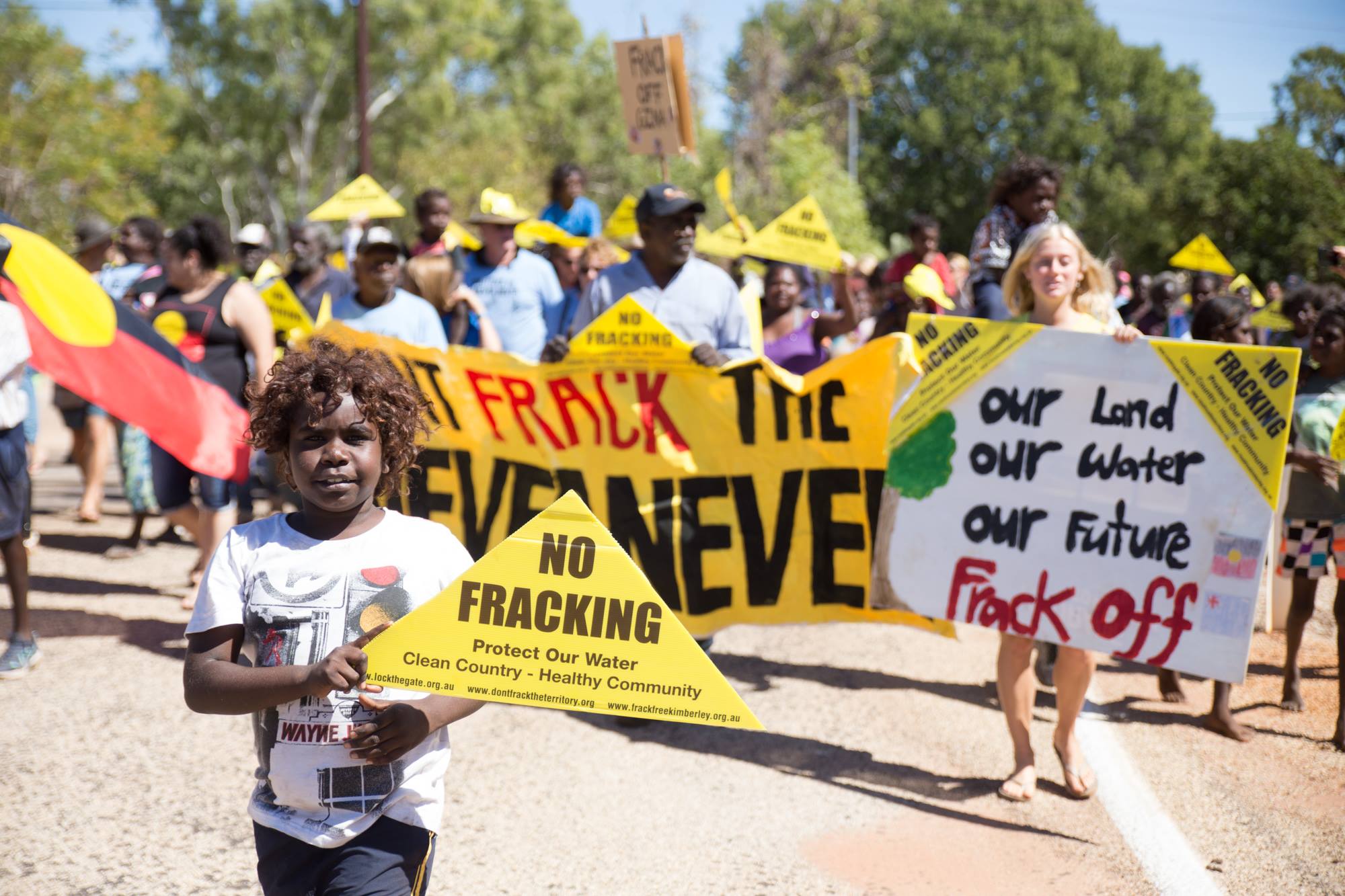 A small boy holds a No Fracking triangle in the foreground and the background is a group of people holding an anti-fracking banner