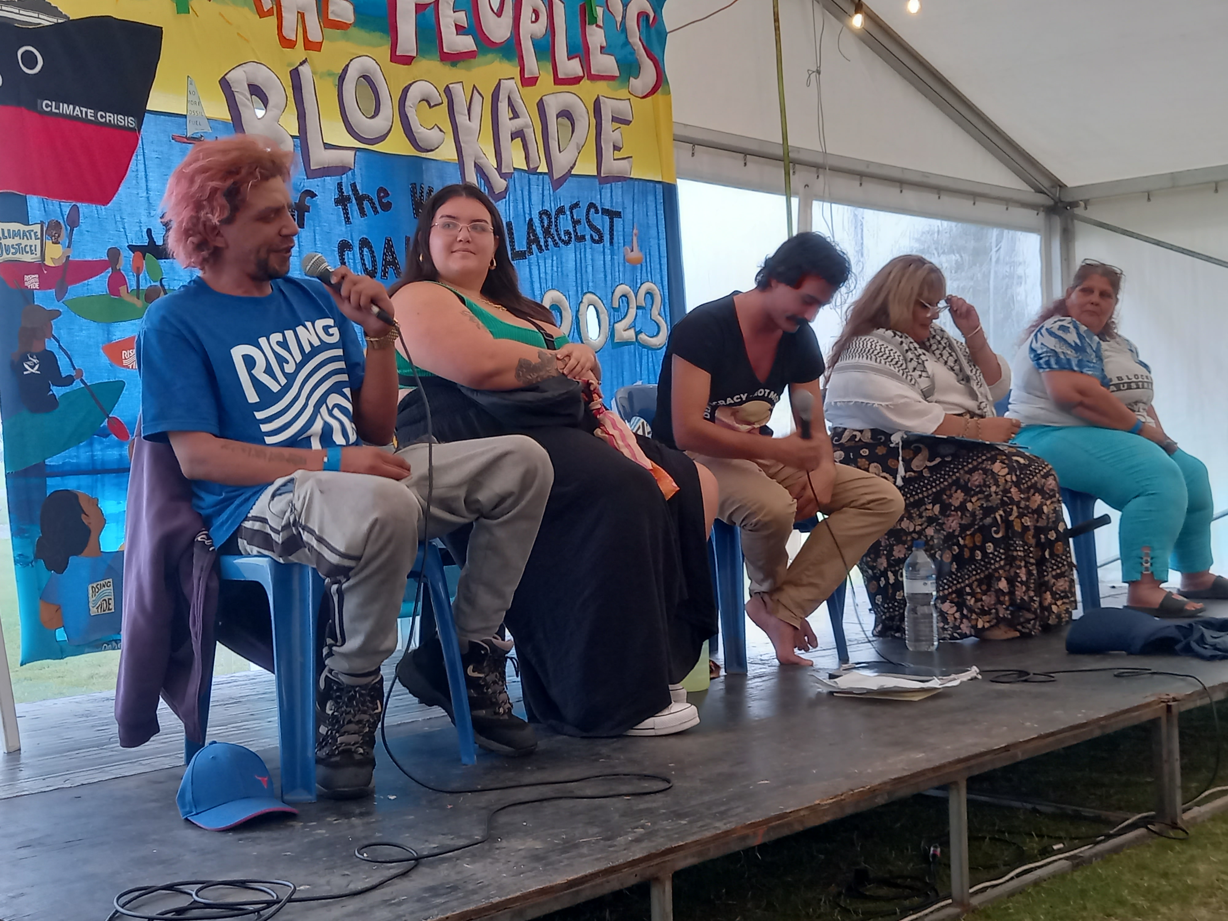 Five first Nations people sitting on chairs on a stage in a large tent with microphones: smilling.