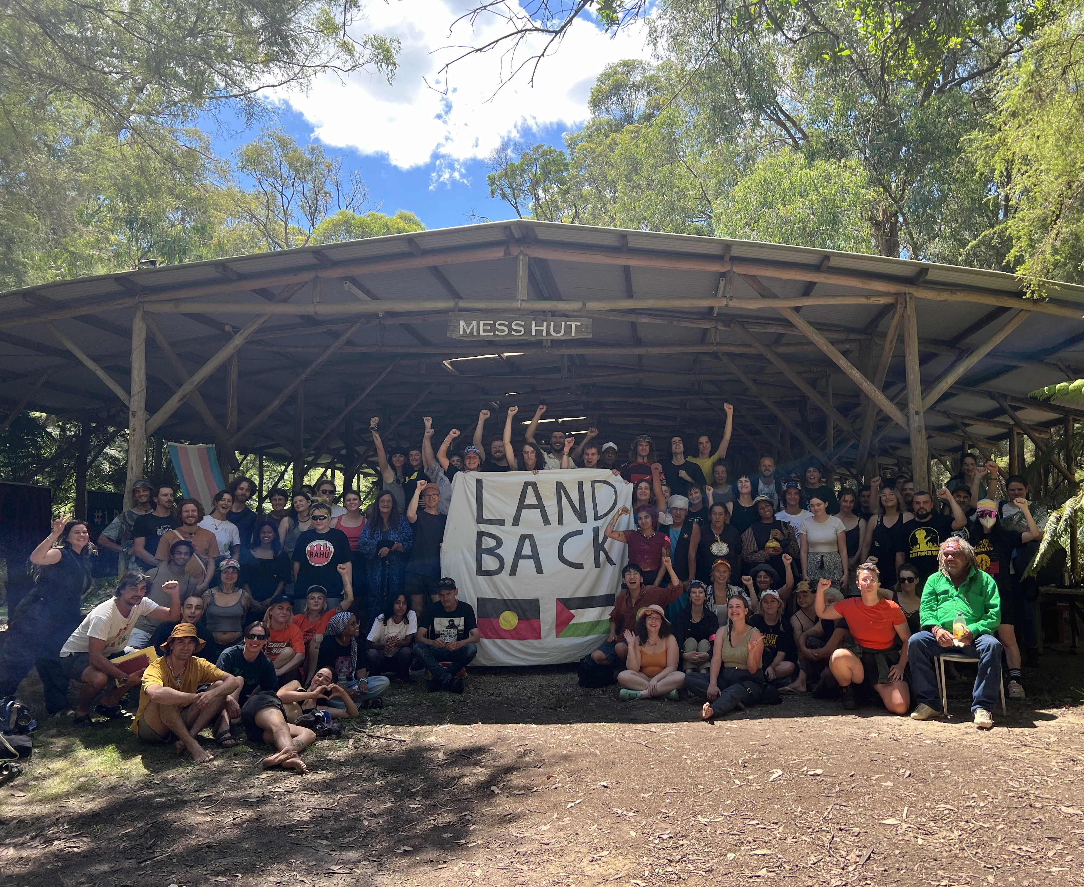 A large group of people crowded at the entrance of an open-walled mess hut at ASEN 2024 Training Camp. They're gathered around a banner that says Land Back and has the Aboriginal and Palestinian flags painted on it.