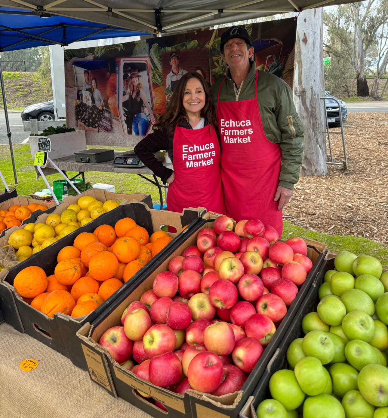 Image of two stallholders (a man and a woman) at the Echuca Farmers' Market standing at their stall. They are selling green and red apples, oranges and lemons. Both stall holders are wearing bright red aprons bearing the words 'Echuca Farmers' Market'.