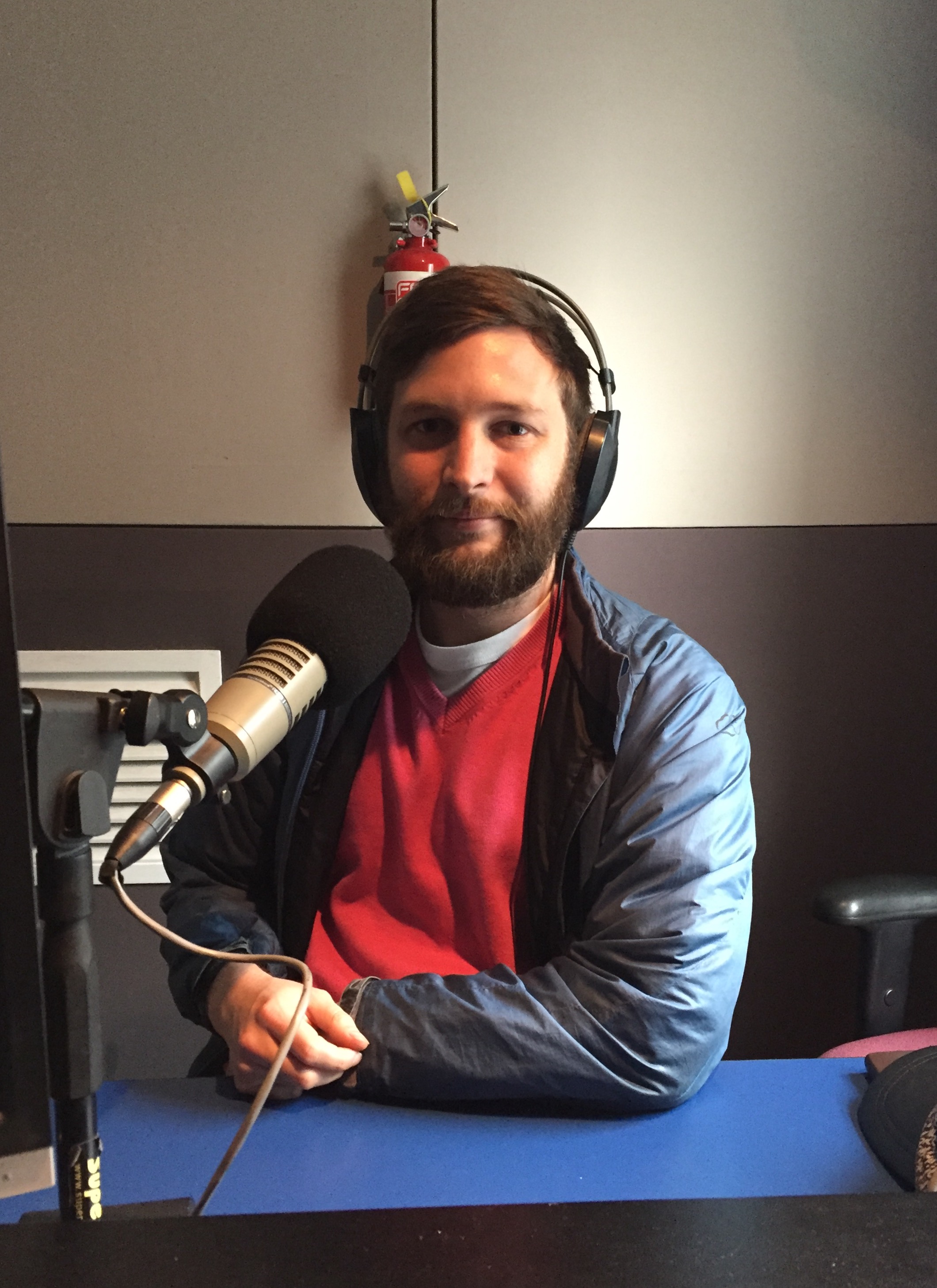 Pat Simons. White male, seated, facing the camera, pictured from the chest up. He is in a recording studio, wearing headphones and there is a microphone on the desk in front of him. He's wearing a pink jumper and a blue vinyl coat.