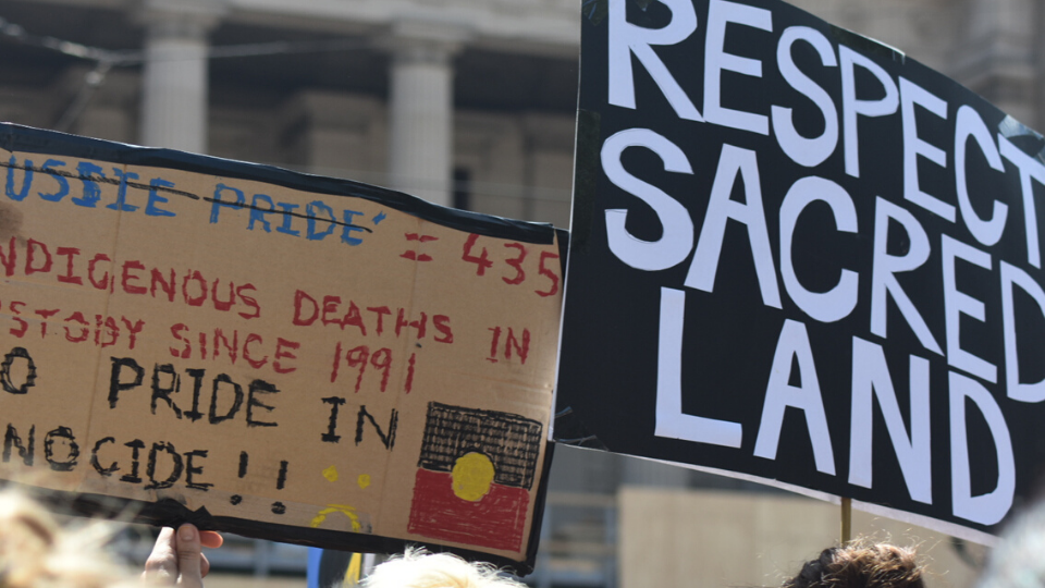 rally placards saying "respect sacred land" and "435 Indigenous deaths in custody since 1991: No pride in genocide"