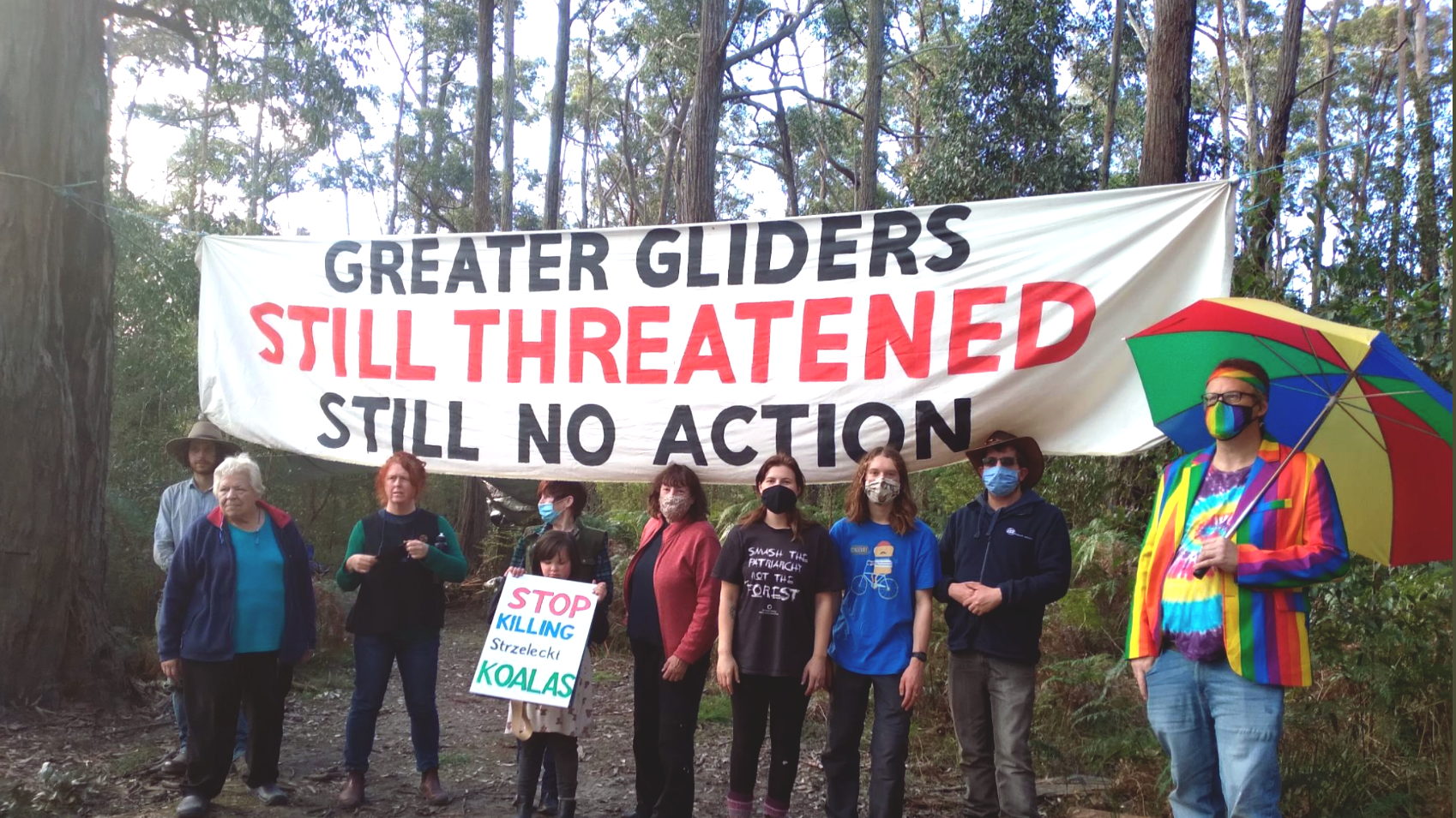 eight activists stand in front of a forest copse at the Alberton West Forest blockade holding a banner that says ‘greater gliders still threatened—still no action’. A small child stands in front of them holding a sign that says ‘stop killing Strzelecki koalas’. A tall middle aged man wearing rainbow coloured clothes and holding an open rainbow striped umbrella stands to the side looking fabulous.