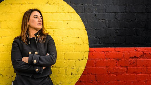 Lidia Thorpe standing in front of wall with Aboriginal flag painted on it looking very staunch