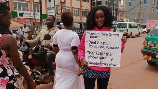 Vanessa Nakate is standing on the street holding a protest sign that says, 'Green love, green peace; beat plastic, polyethylene, pollution; thanks for the global warming; climate strike now.' in the background are people walking by, traffic & buildings.