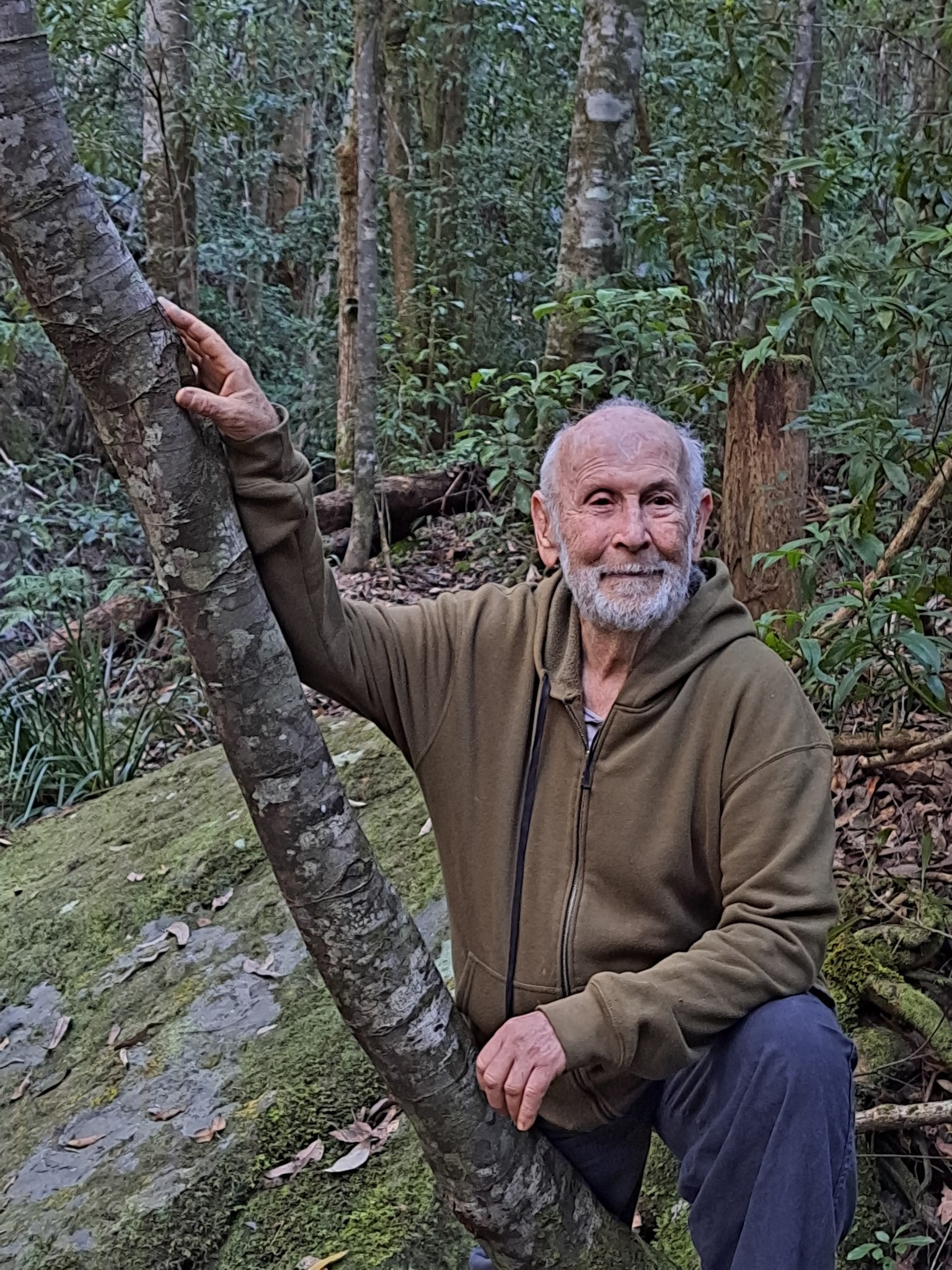 An older man with his hand on a tree trunk