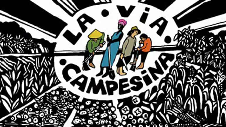  La Via Campesina banner artwork featuring a mandala style blank & white panel with illustrated cartoon style sections of corn, sun flowers, squash and other food cops growing all pointing towards an elliptic/egg-shaped centre featuring for peasant workers from around the world holding/using different farming tools. Their skin and clothes are the only colourful objects in the graphic. They are framed by the words La.Via.Campesina.