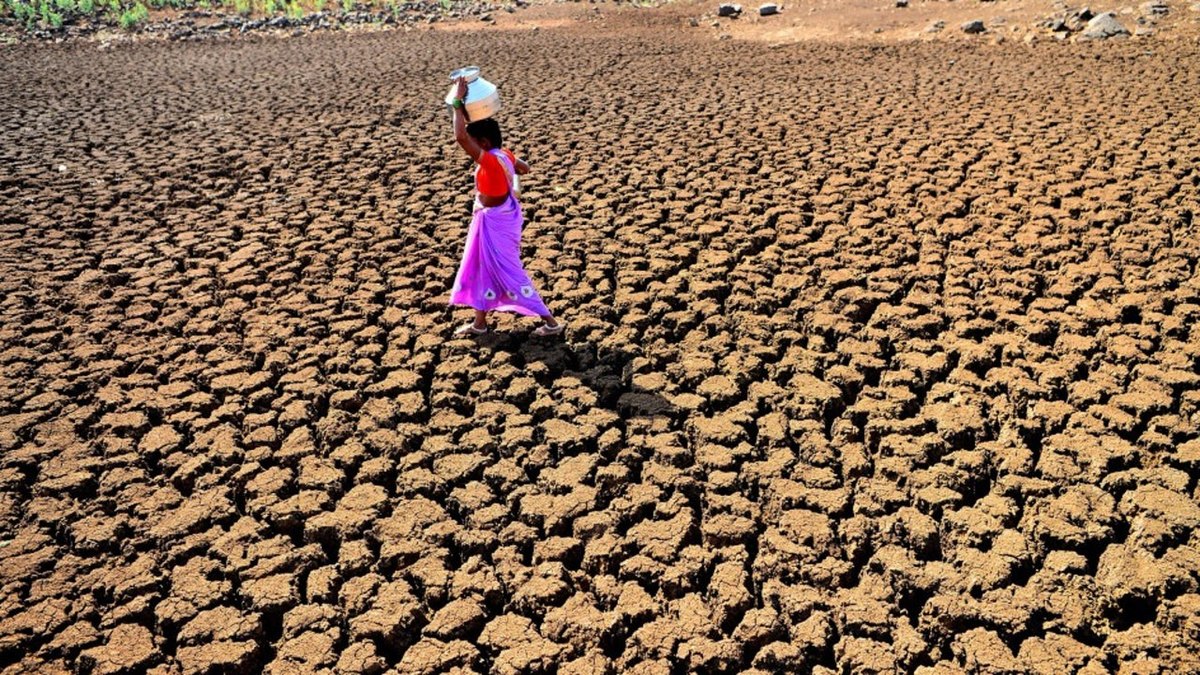 Woman dressed in brightly coloured sari carrying a jug of water on her head and walking across very parched ground