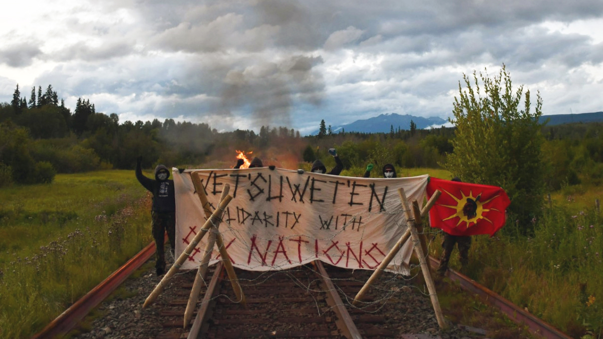 Several anonymous masked protesters wearing black pants, black hoodies, gloves and dark sunglasses stand defiantly with fists in the air at a barricade that has been erected across train tracks in a remote area. One of the protesters is holding up a Wet'suwet'en flag, which has a red background with a yellow sun, and an image of the profile of a Wet'suwet'en warrior wearing headdress in the centre of the sun. The barricade is constructed with 2 large, solid wooden tripods across tracks and a large banner st