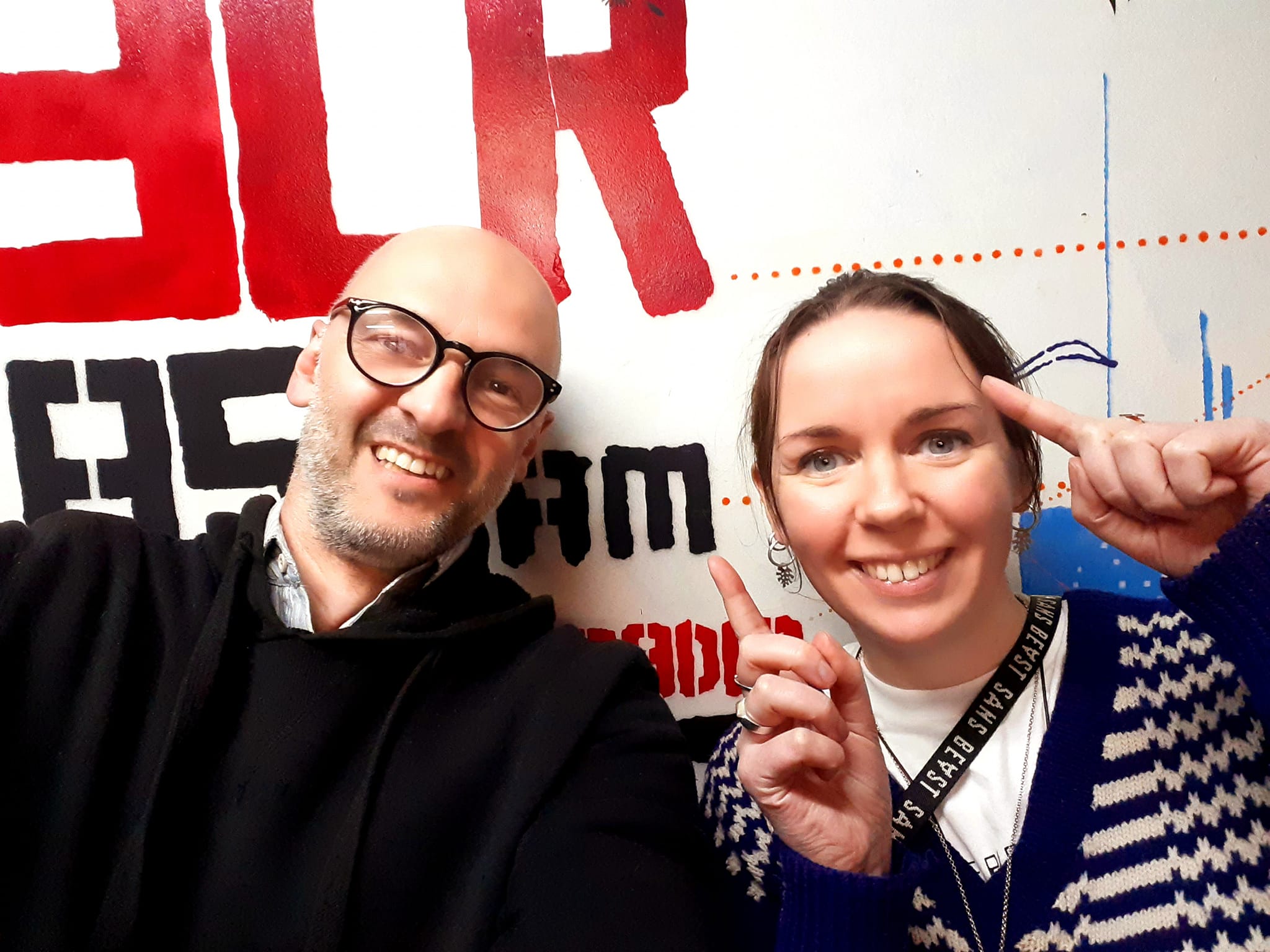 Photograph of presenters Brendan Bonsack (Left) and Carmen Main (Right), smiling at the camera. Behind them is a partial wall mural with the 3CR Radio logo.