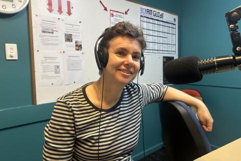 A woman with short hair and wearing a striped top sits smiling and looking at the camera. She has one arm leaning on a chair back and is wearing headphones. 