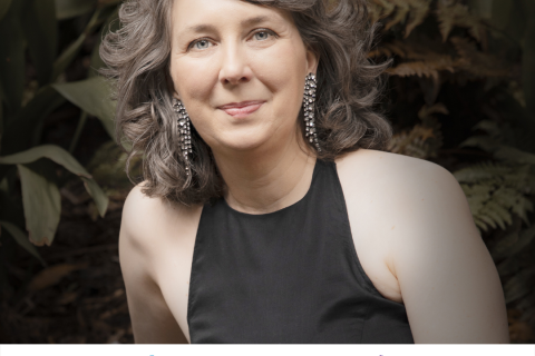 A caucasian woman with brown and grey shoulder length wavy hair in a black halter top smiles at the camera, she is against an outdoor backdrop of green plants and leaves 