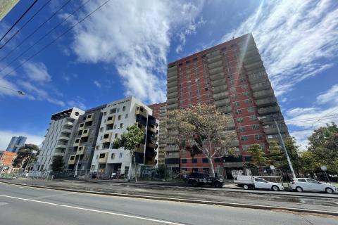 New ‘community’ housing currently being built alongside the decommissioned public housing estate at 141 Nicholson St, Carlton. The new building on the left is short with stainless steel cladding and no green space, and the boarded up 16-storey brick building on the right has balconies and graffiti on the base of its façade. The sky is cloudy and traffic is passing on a street in the foreground with tram tracks and multiple lanes.