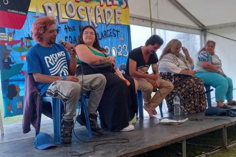 Five first Nations people sitting on chairs on a stage in a large tent with microphones: smilling.