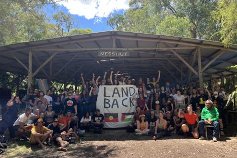 A large group of people crowded at the entrance of an open-walled mess hut at ASEN 2024 Training Camp. They're gathered around a banner that says Land Back and has the Aboriginal and Palestinian flags painted on it.