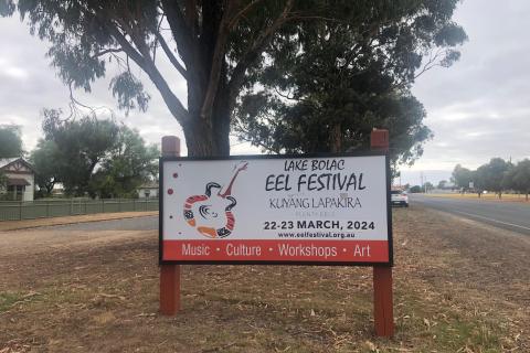 Sign reading 'Lake Bolac Eel Festival - Kuyang Lapakira - 22- 23 March, 2024'.  Sitting on a grassy strip in the middle of two roads with gum trees behind.