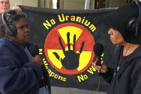 3CR Presenter Viv Malo speaking with Aunty Vicki Abdullah (Tjiwarl Traditional Owner & co-chair of the Australian Nuclear free Alliance at  the IMARC blockade in Melbourne on Oct 28. They are facing each other wearing headphones and holding microphones standing in front of a banner that says "no uranium, no weapons, no way".