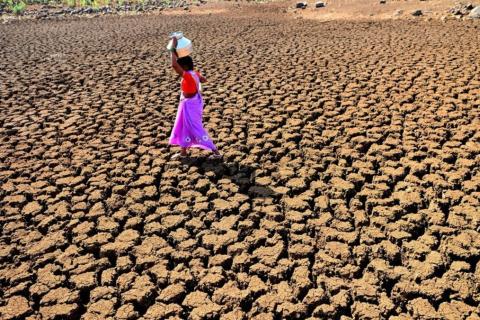 Woman dressed in brightly coloured sari carrying a jug of water on her head and walking across very parched ground