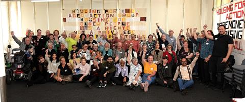 Dozens of HAAG members and staff stand in front of a quilted banner, their first raised in the air. The banner reads Housing for the Aged Action Group, housing for the aged is a right not a privilege.